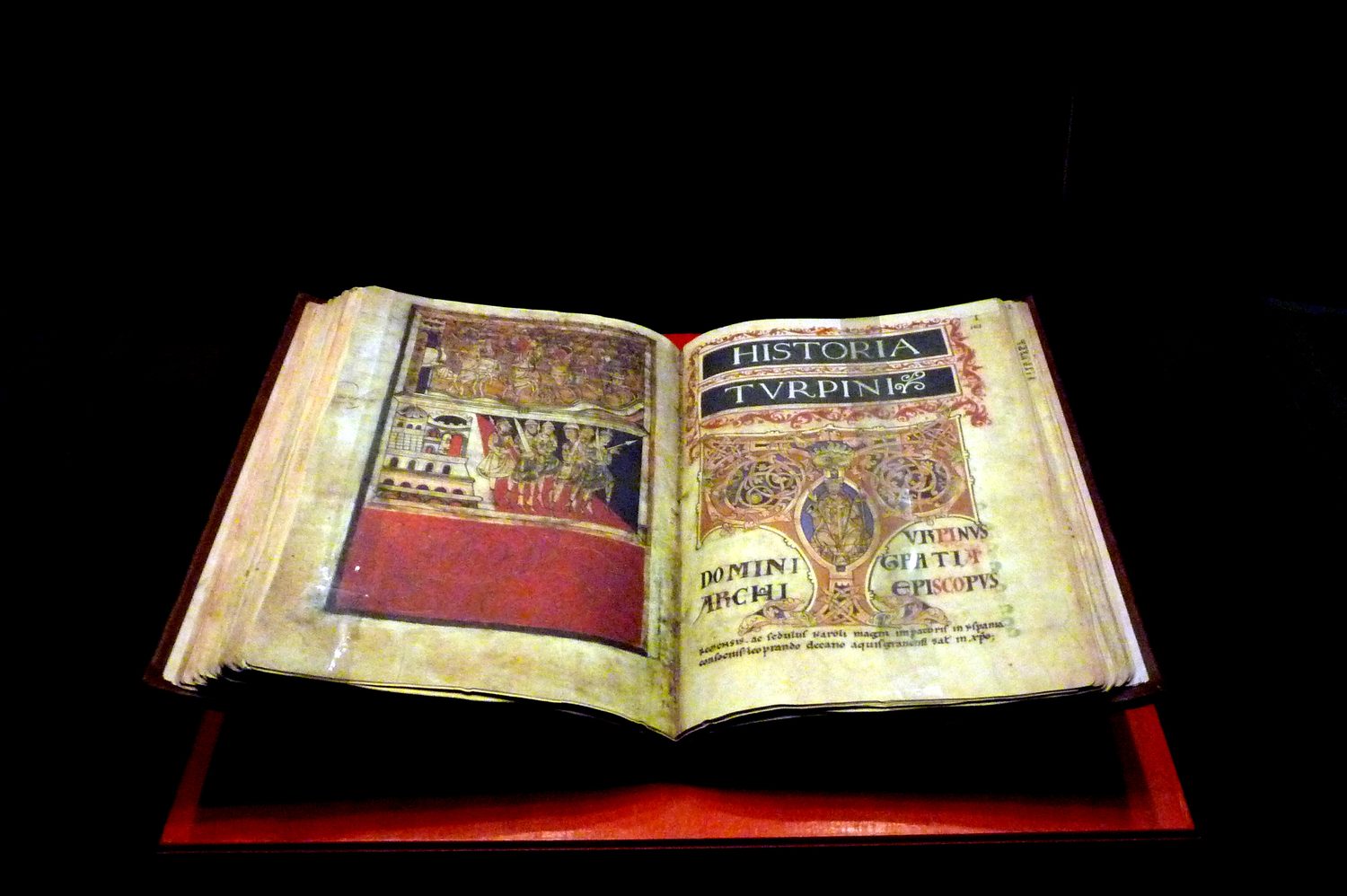 O Codex Calixtinus aberto numa vitrine, mostrando a página iluminada da Historia Turpini, com cenas de Carlos Magno e iniciais decoradas