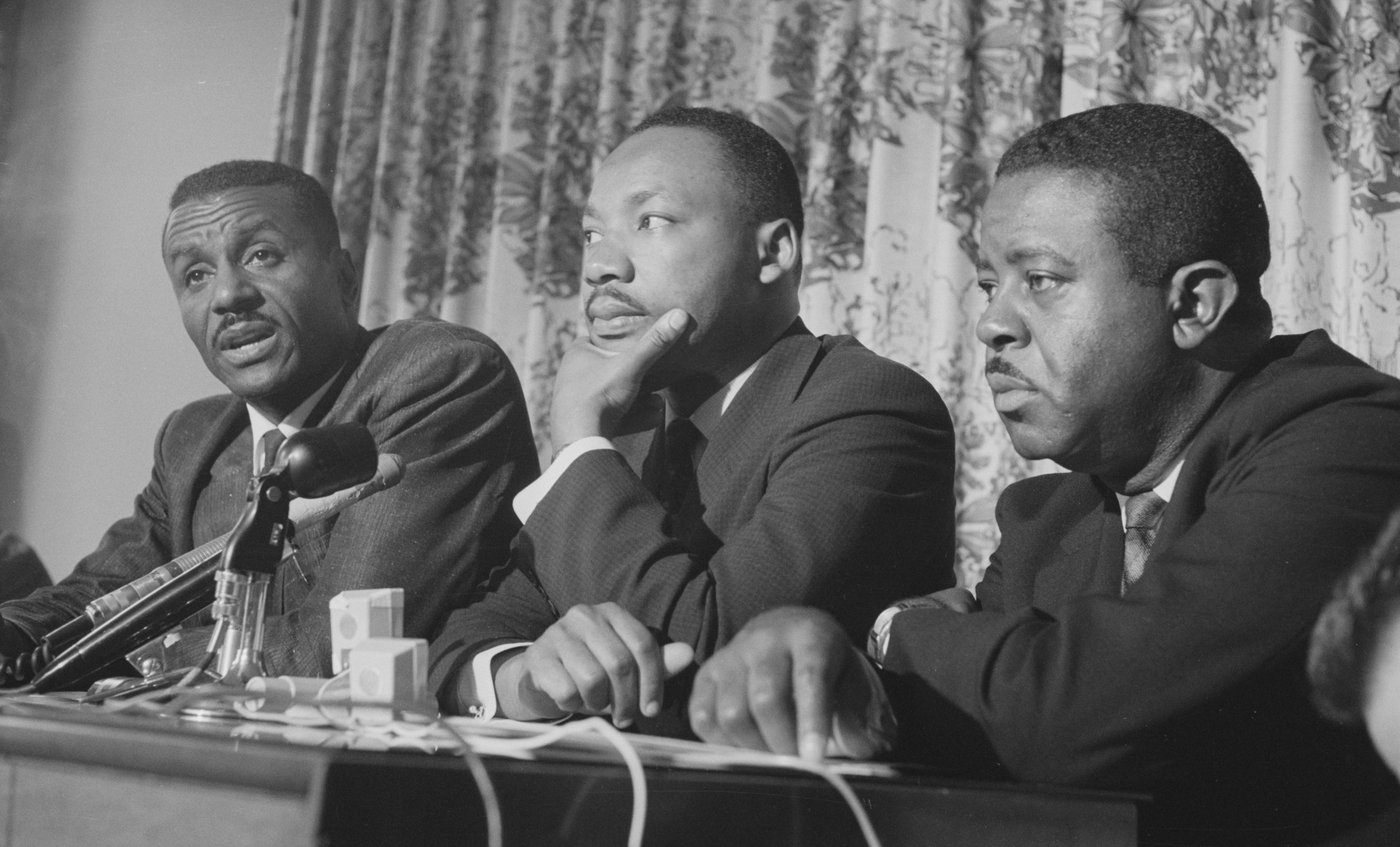Fred Shuttlesworth, Martin Luther King Jr. e Ralph Abernathy sentados lado a lado diante de microfones, durante uma coletiva de imprensa em Birmingham
