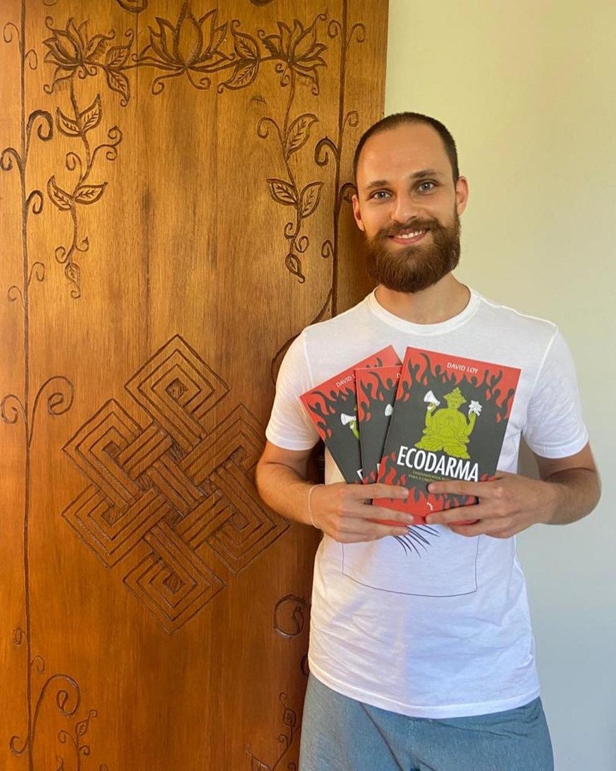 Lucas Ghisleni sorrindo, segurando três exemplares do livro Ecodarma, de David Loy, ao lado de uma porta de madeira entalhada com flores de lótus