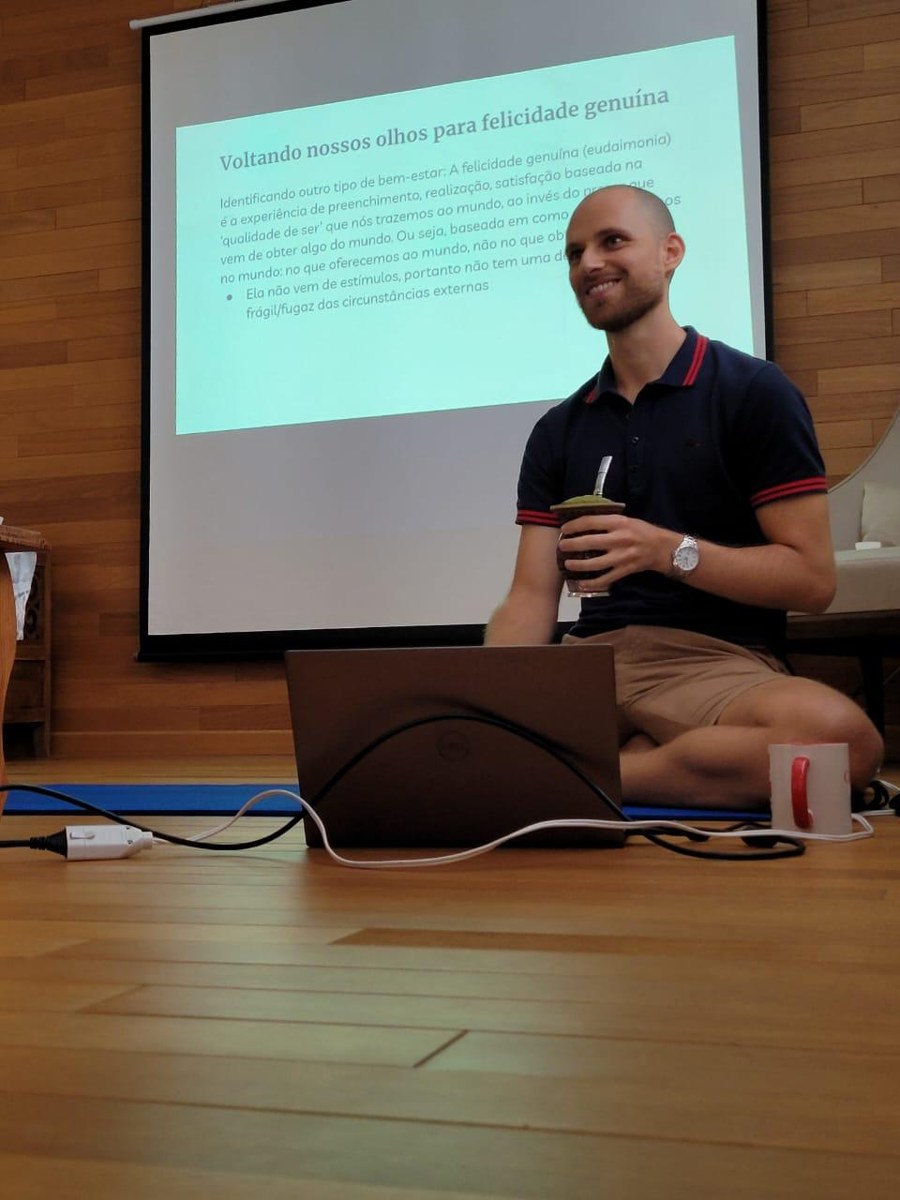 Lucas Ghisleni sentado no chão, sorrindo e segurando uma cuia de chimarrão, diante de um telão com um slide sobre felicidade genuína