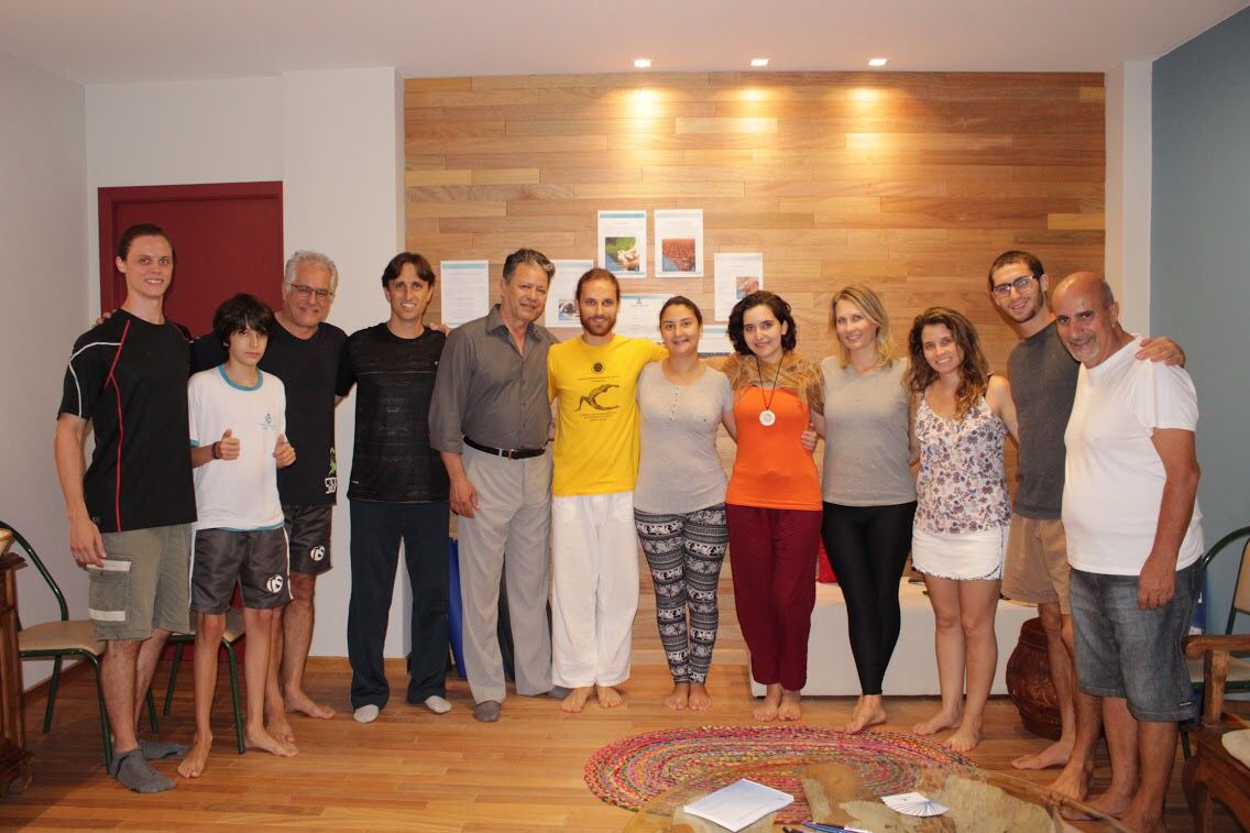 Foto de grupo numa sala com parede de madeira — Lucas de camiseta amarela ao centro, cercado pelos primeiros alunos, com um tapete redondo no chão