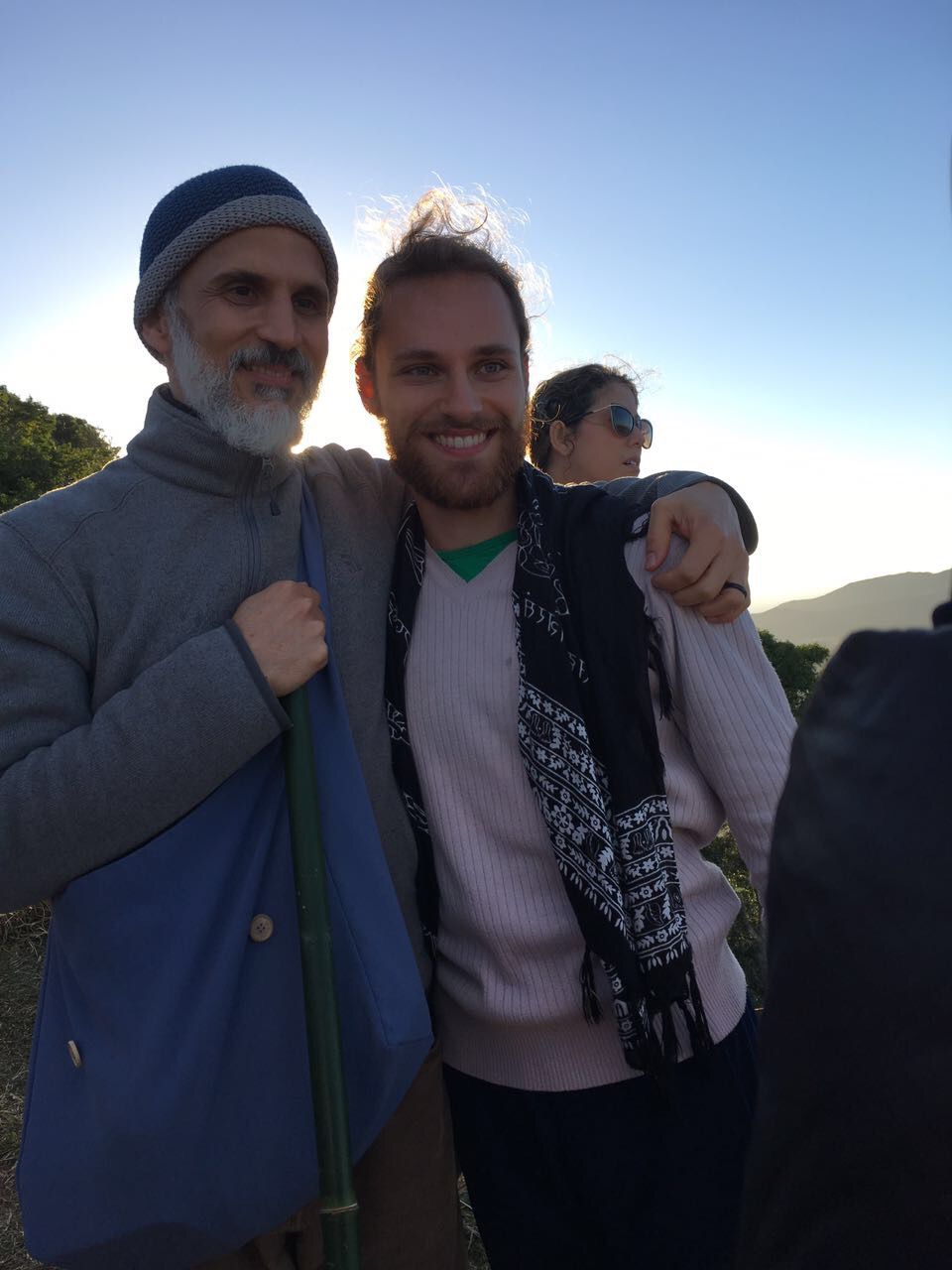 Lucas ao lado de Marco Schultz, um homem de barba grisalha e gorro de tricô, num morro ao entardecer