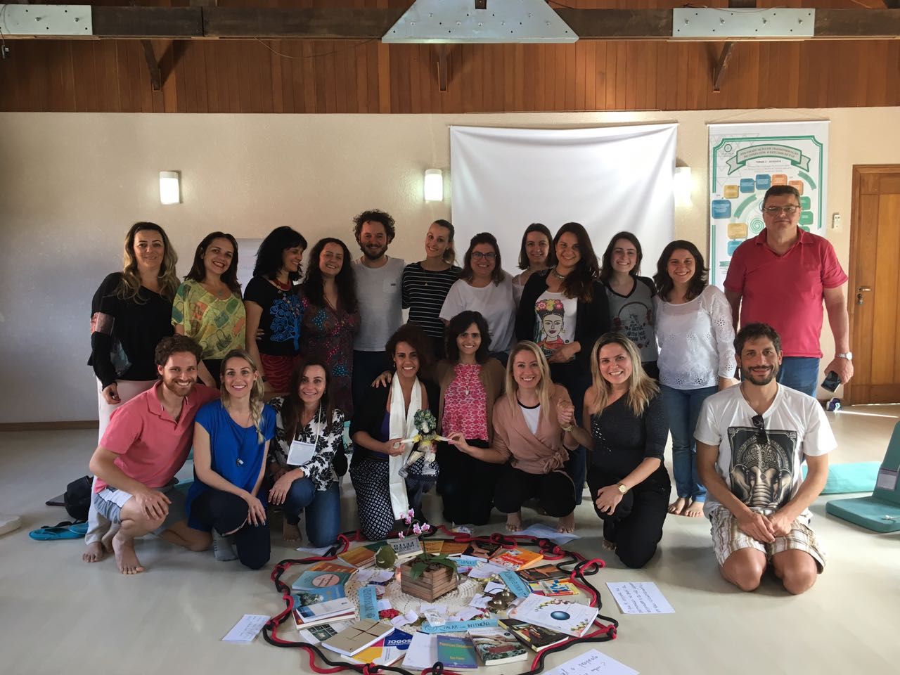 Foto da turma da pós-graduação — cerca de vinte pessoas reunidas numa sala de retiro, com uma mandala de livros e objetos no chão e o cartaz do curso ao fundo